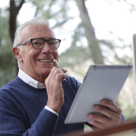 Photo of an elderly man. He is sitting by a window, in front of a computer, reading from a pad of paper