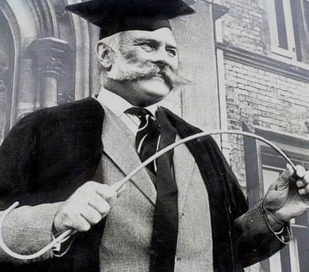 Photo, looks like a still from an old movie, showing an old-style teacher complete with gown and cane.