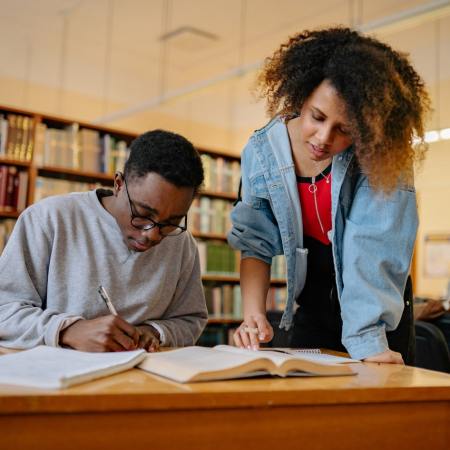 A pair of people. One is sitting down, writing. The other is standing over his shoulder, looking at what is being written.